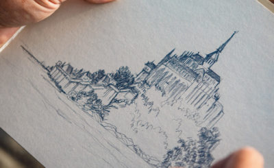 Artiste dessinant le Mont-Saint-Michel au crayon, focus sur ses mains et son dessin