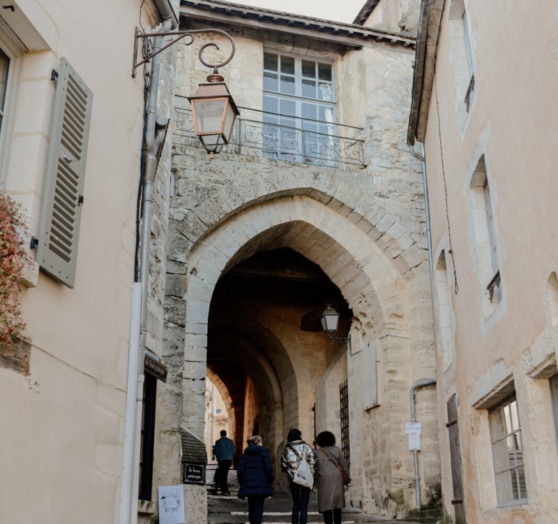 Rue avec porche en pierres, un groupe de personne marche.