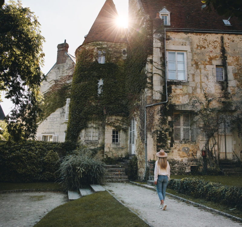 Rue de Mortagne-au-Perche, le soleil tape derrière une tour recouverte de vigne. Une femme avec un chapeau se dirige en sa direction.