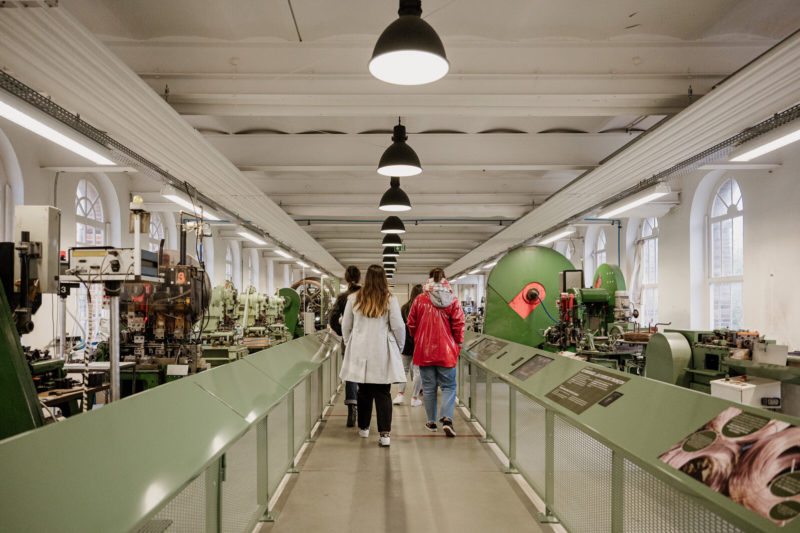 Personnes visitant la Manufacture Bohin. L'allée est bordée de machines et de panneaux explicatifs
