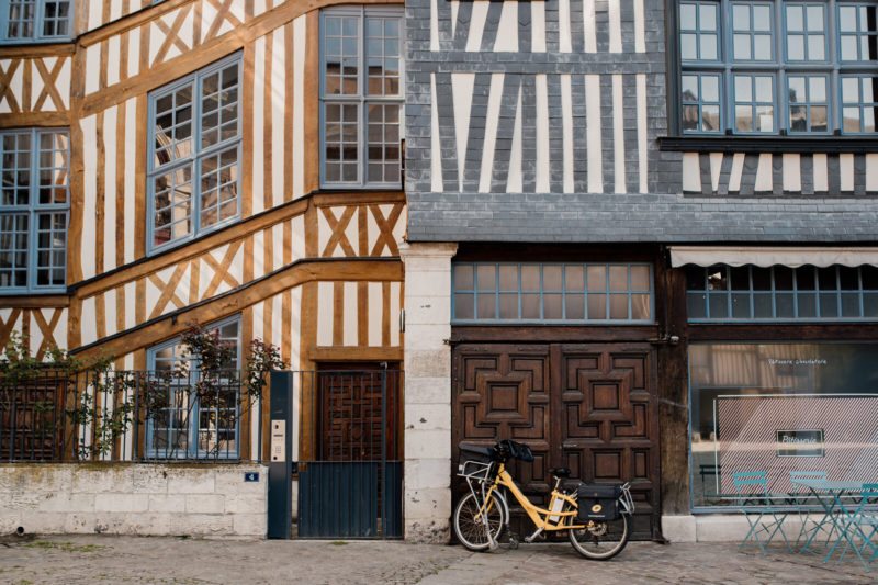 Façades mitoyennes en colombages, typiques rouennaises, à gauche de couleur marron claire et à droite grise, avec une porte en bois et un vélo jaune placé devant.