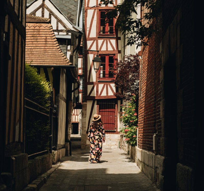 Rue du Chanoines à Rouen. Rue étroite avec maison en colombage rouge au fond, une femme vêtue d'une robe et d'un chapeau s'y dirige.