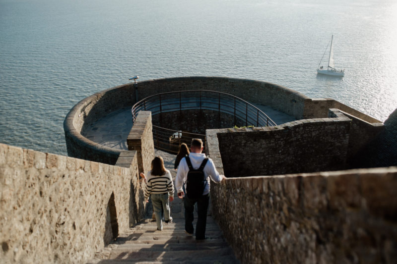 Remparts du Mont-Saint-Michel, une femme et un homme descendent les escaliers. La prise de vue de haut permet d'observer les remparts et la baie.